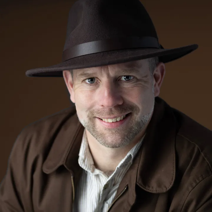 Antony Butts in a brown felt fedora and field jacket, smiling at the camera