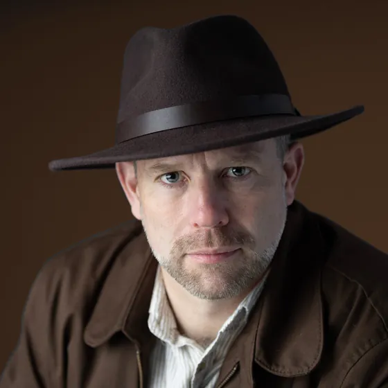 Antony Butts in a fedora and field jacket, looking straight at the camera