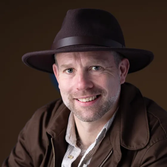 Antony Butts in a fedora and field jacket, head tilted, smiling